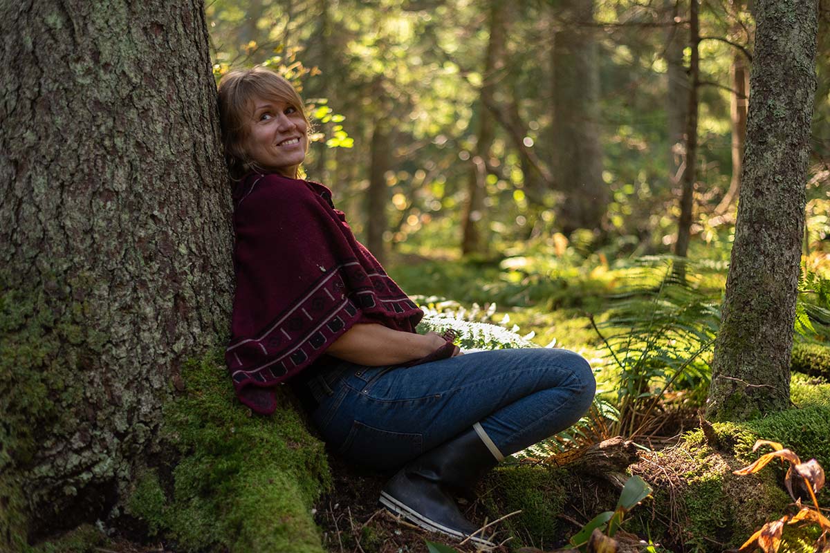 A person in a red shawl and jeans sits against a tree in a sunlit forest, smiling and looking slightly upwards.