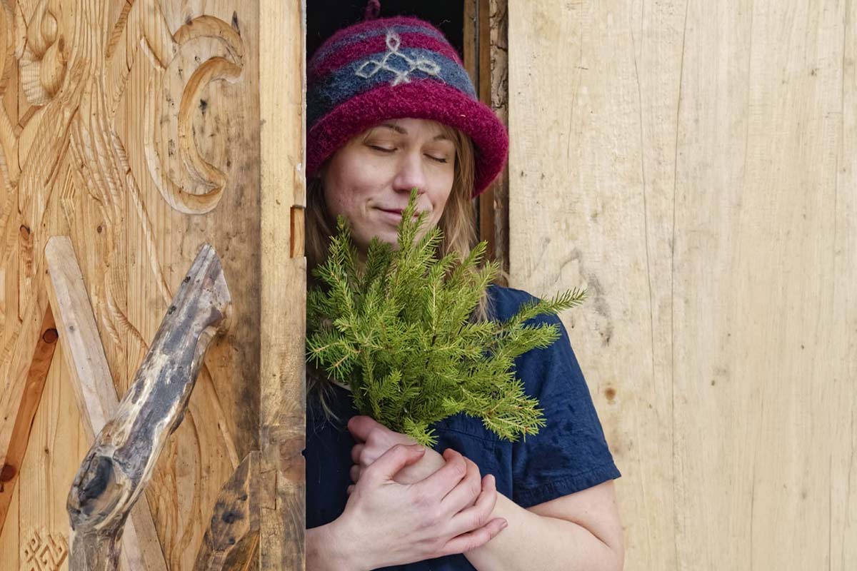 A person in a red hat stands in a wooden doorway, holding a small green twig and smiling gently with closed eyes.