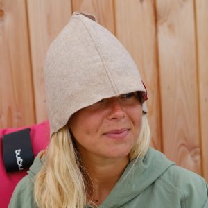 A woman with long blonde hair wears a Sauna Hat that covers her eyes as she sits in front of a wooden wall, paired with a green hoodie.