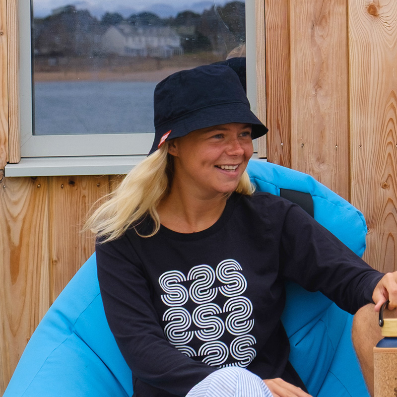 Woman in a black hat and long-sleeved shirt sits on a blue beanbag chair outdoors, smiling, with wooden walls and a window in the background.