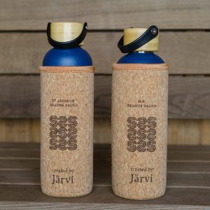 Two blue water bottles with wooden caps and cork sleeves stand side by side on a wooden surface. The sleeves are labelled "SEASIDE SAUNA" and "created by Järvi.