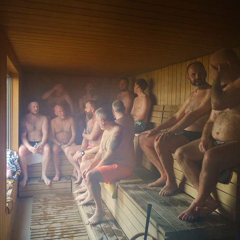 A group of men in swimwear sit and talk inside a wooden sauna room, with some looking out the window and others engaged in conversation.