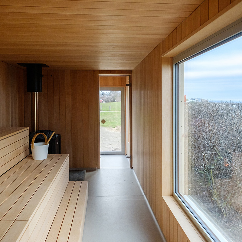 Interior of a modern wooden sauna with a bench, bucket, stove, and large window overlooking outdoor trees and sky.