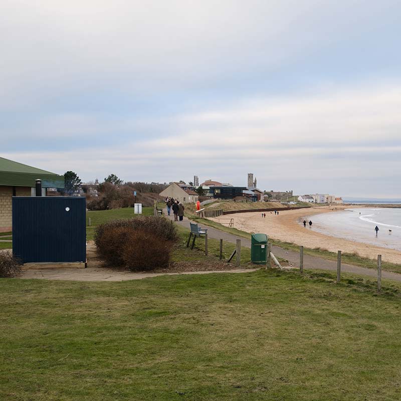 People walk along a paved path by a sandy beach with grassy areas, a blue shed, and buildings in the distance under a cloudy sky.