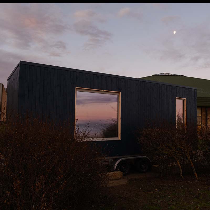A small, dark-coloured tiny house on wheels sits among bushes at dusk, with large windows reflecting the sky and a faint moon visible above.