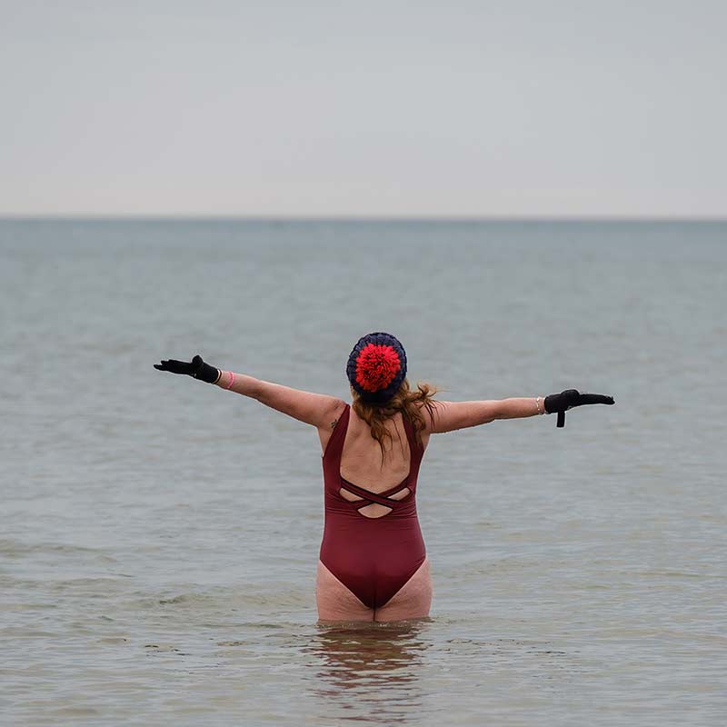 Person wearing a red swimming costume, gloves, and a swimming cap stands waist-deep in calm water with arms outstretched, facing the horizon.