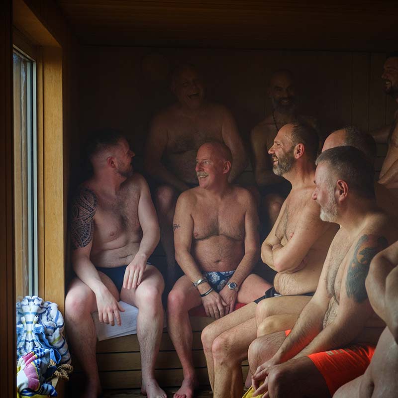 A group of men in swimwear sit closely together, talking and laughing inside a wooden sauna with a window on the left.