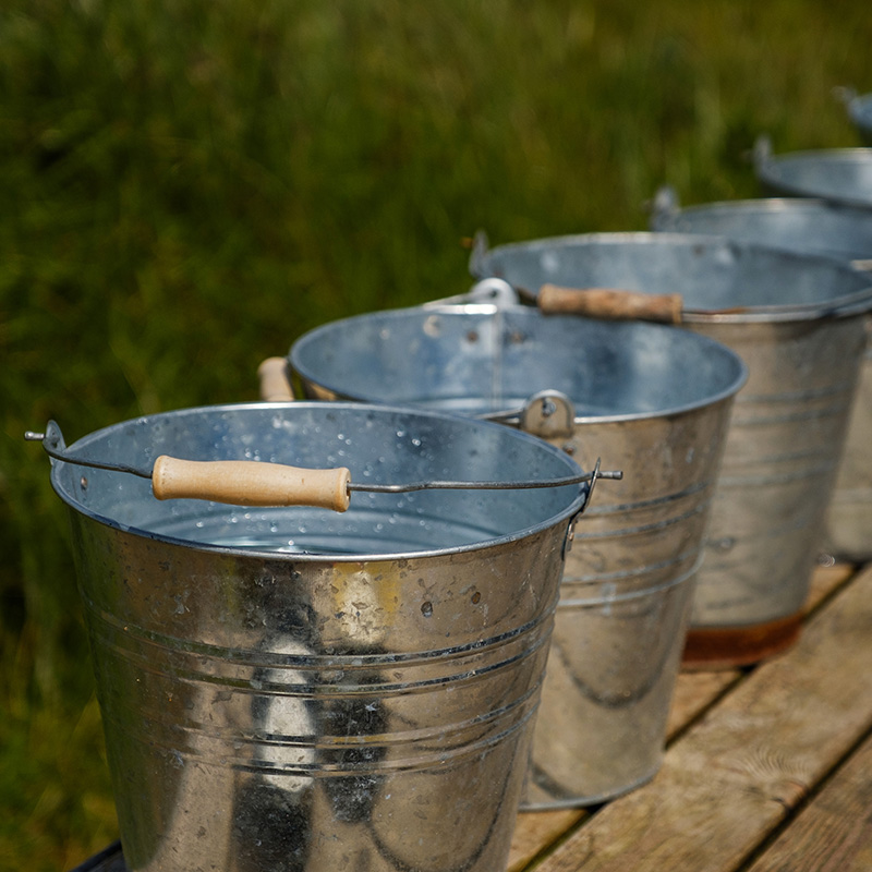 Scottish-Seaside-Saunas-Elie-09 A row of shiny metal buckets with wooden handles is lined up on a wooden surface outdoors, with grass in the background.