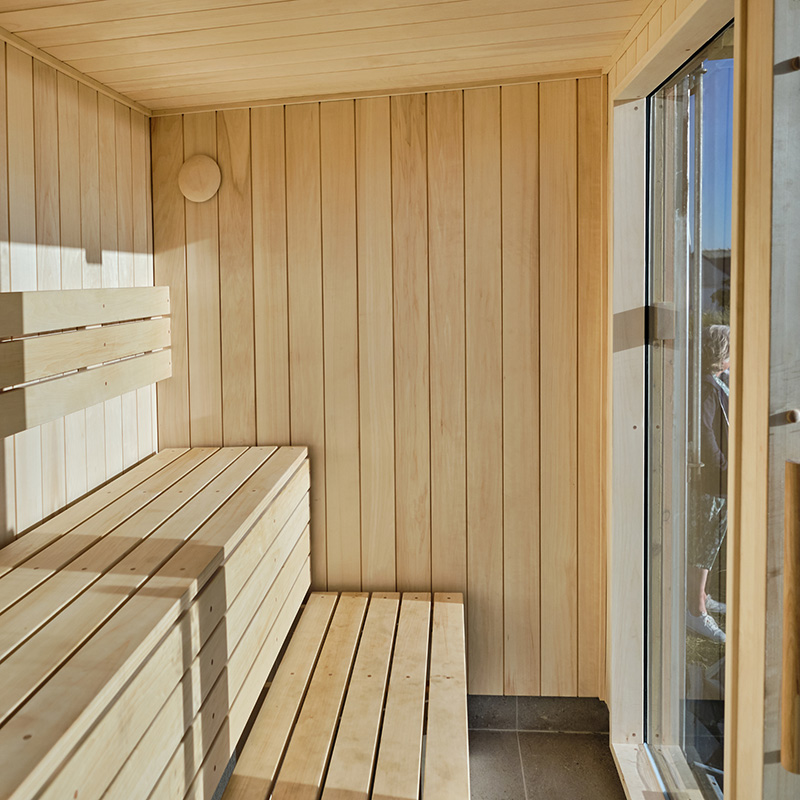 Scottish-Seaside-Saunas-Elie-10 Interior of a modern wooden sauna with bench seating, light wood panelling, and a glass door letting in sunlight.