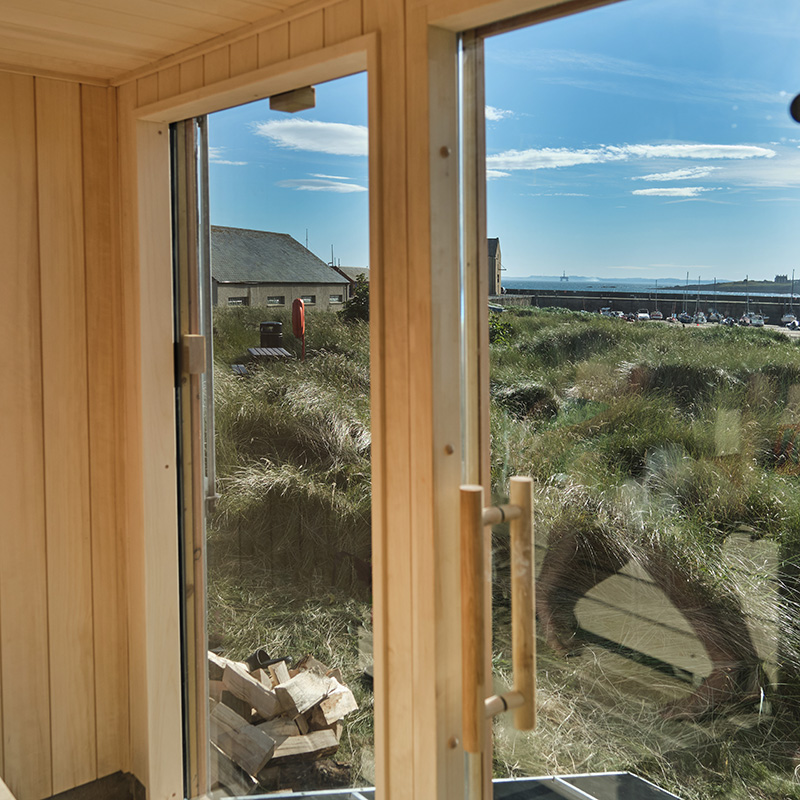 Scottish-Seaside-Saunas-Elie-11 View through a glass door in a wooden interior, showing grassy dunes, a stack of firewood, houses, and a clear blue sky outside.