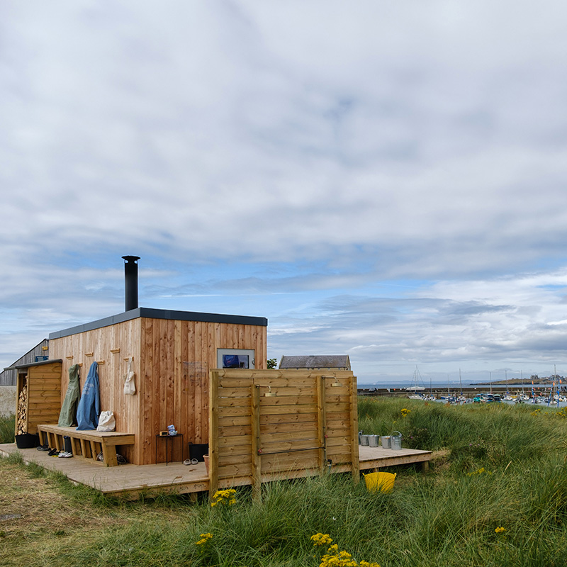 Scottish-Seaside-Saunas-Elie-12 Small wooden cabin with an outdoor shower, benches, and hanging towels, set on grassy land near a marina under a cloudy sky.