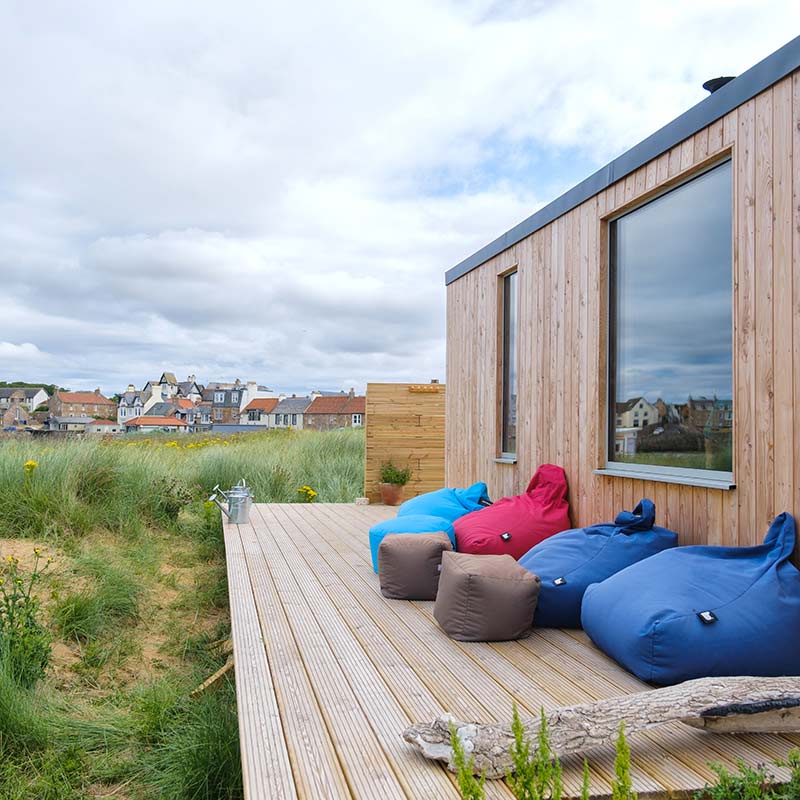 Scottish-Seaside-Saunas-Elie-13 Wooden decking with colourful bean bags next to a modern house, overlooking grassy dunes and a distant view of a village under a cloudy sky.