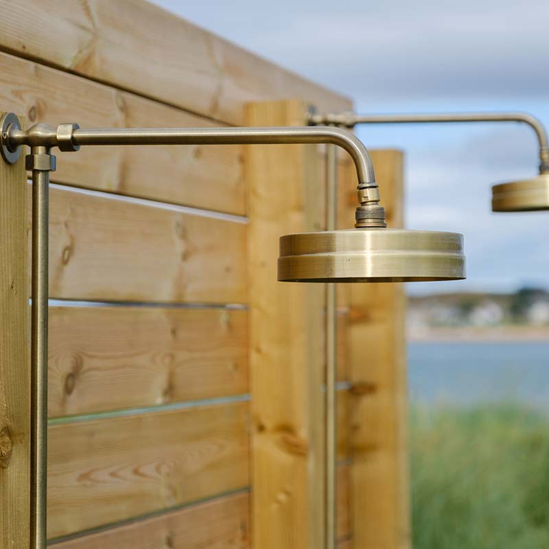 Scottish-Seaside-Saunas-Elie-14 Close-up of a brass outdoor shower fitting mounted on a wooden privacy screen, with blurred natural scenery in the background.