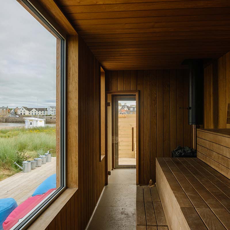 Scottish-Seaside-Saunas-Elie-15 Interior of a wooden sauna with a large window and glass door, overlooking an outdoor landscape with grass, buildings, and a cloudy sky.