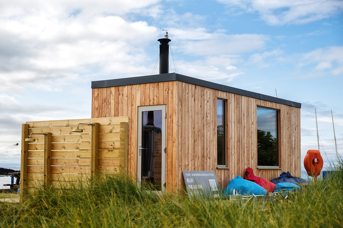 Scottish-Seaside-Saunas-Elie-16 Small modern wooden cabin with large windows, an outdoor shower area, beanbags, and lifebuoys, situated among tall grass under a partly cloudy sky.
