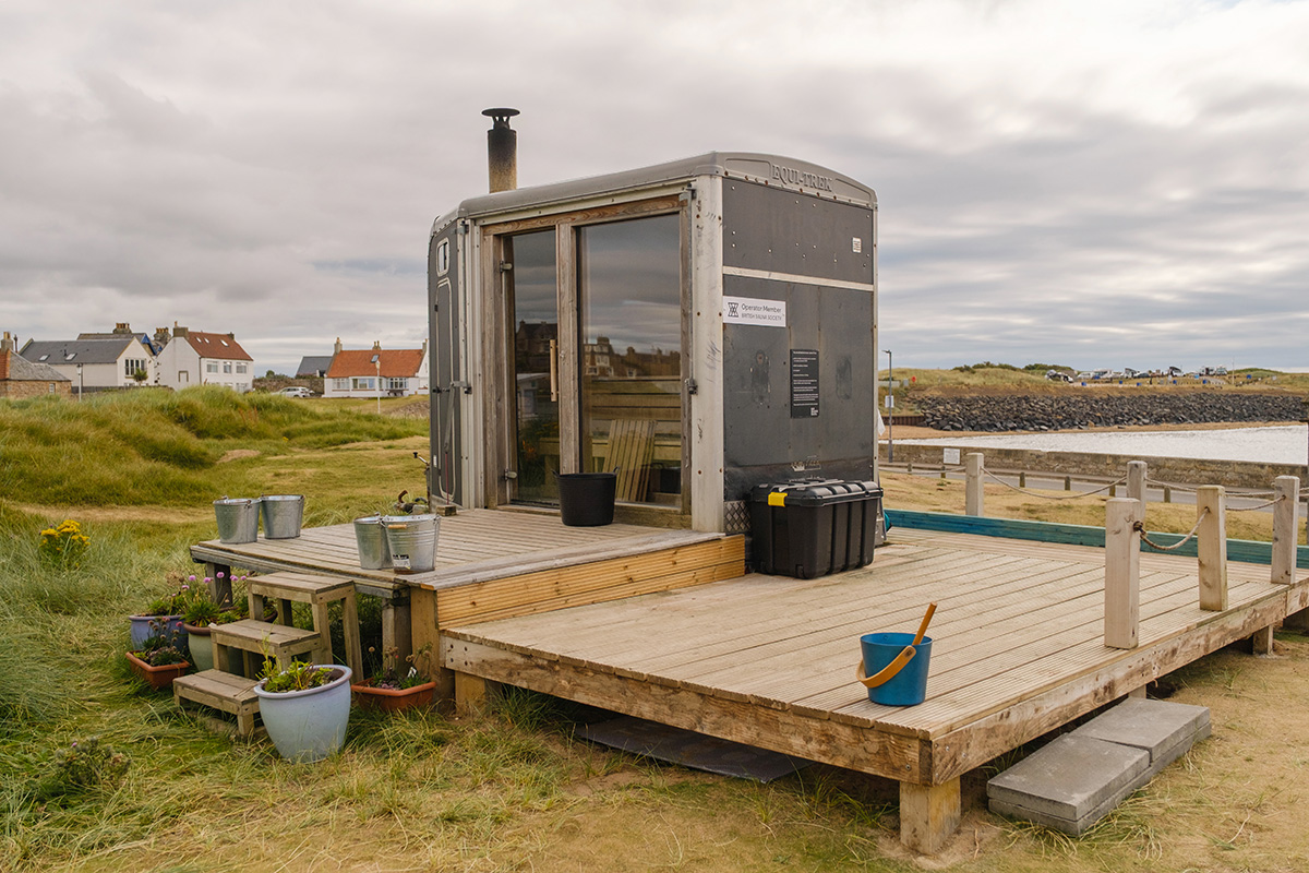 Scottish-Seaside-Saunas-Elie-17 Small modern sauna cabin with glass door sits on a wooden decking near the coast, surrounded by grass, plants, steps, and houses in the background under a cloudy sky.