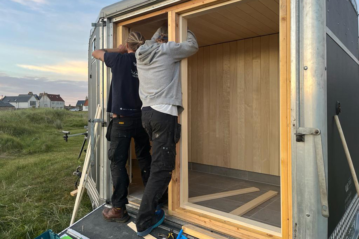 Scottish-Seaside-Saunas-our-story-01 Two people install wooden panelling inside a small structure or caravan set up outdoors near grassy land and houses.