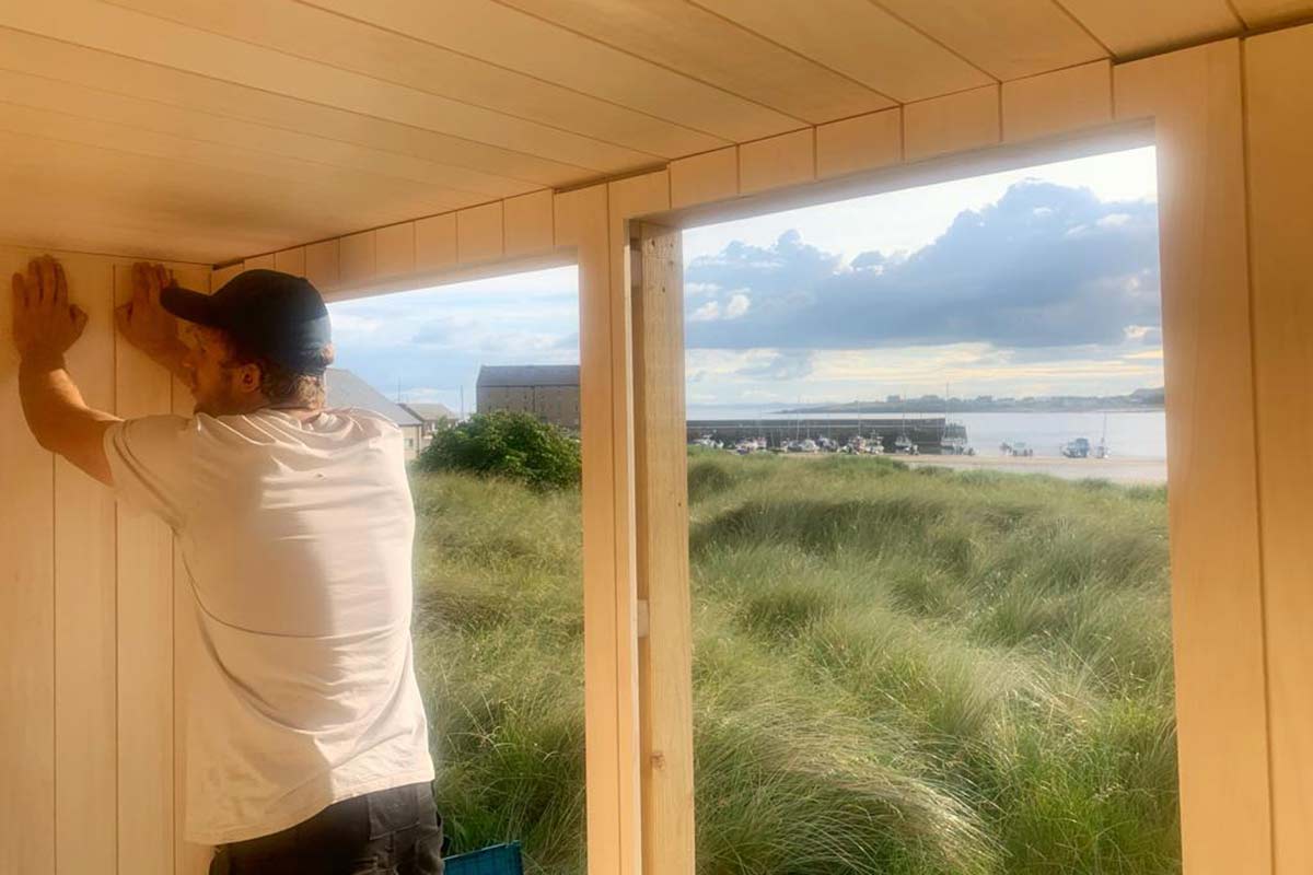 Scottish-Seaside-Saunas-our-story-02 A person in a white shirt and cap installs wooden panelling inside a structure with large windows overlooking grassy dunes and water with boats.