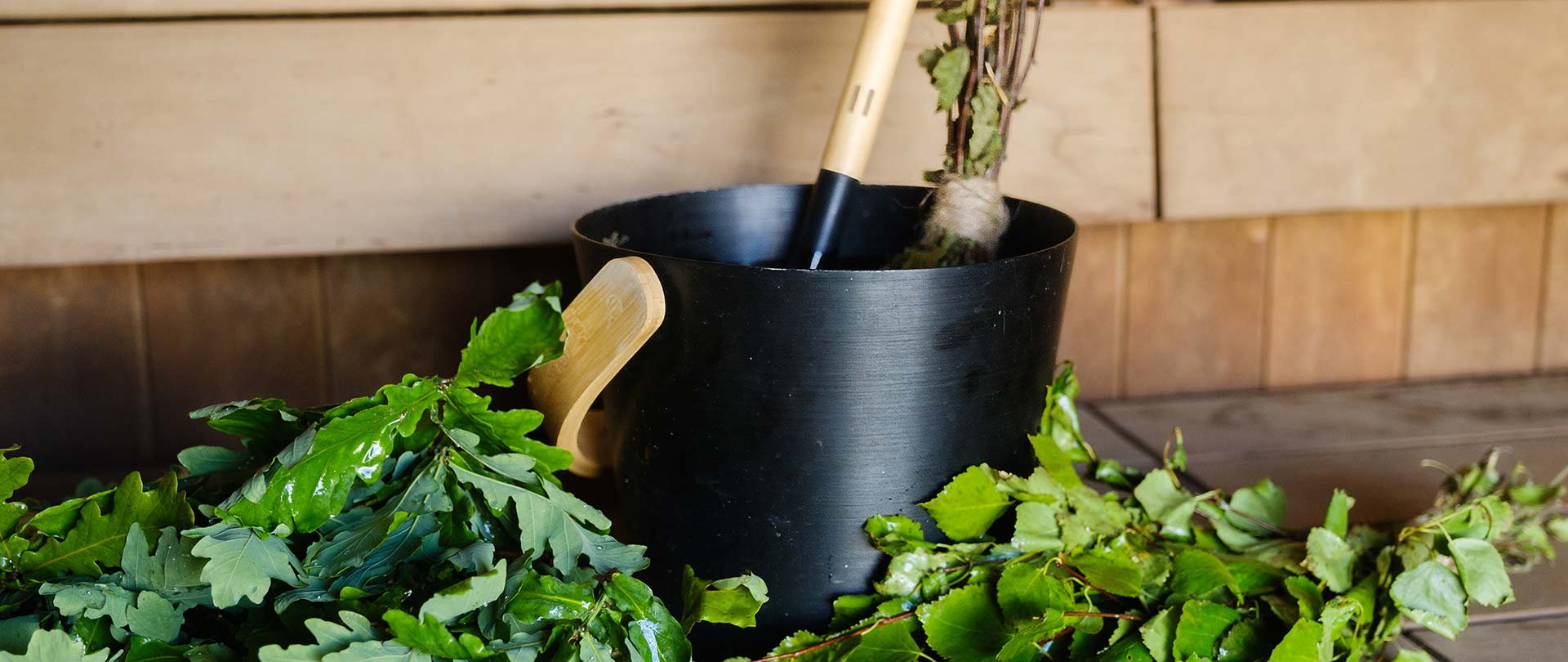A black bucket with a wooden handle, leafy branches, and a wooden ladle sits on a wooden sauna bench.