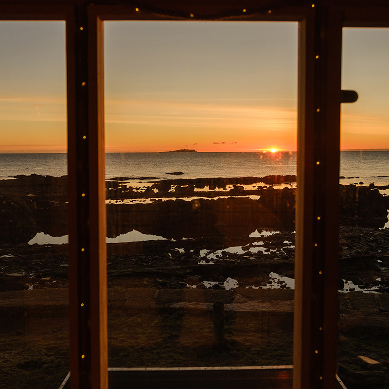 Scottish-seaside-saunas-Cellardyke-01 A sunset over a rocky shoreline is seen through a window with fairy lights, with calm sea and an island in the distance.