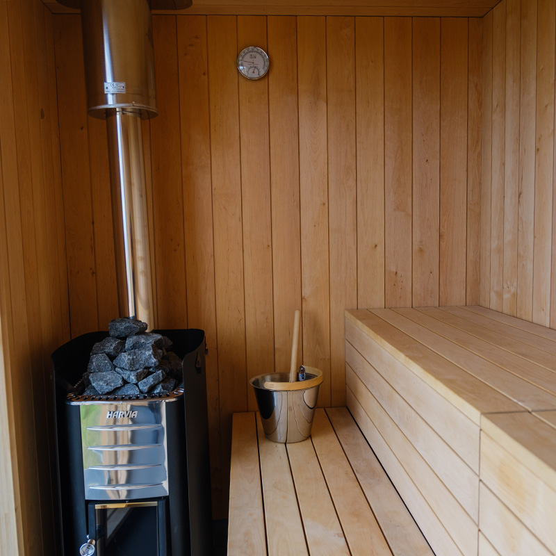 Scottish-seaside-saunas-Cellardyke-02 Wood-panelled sauna room with a metal stove holding sauna stones, a bucket with a ladle, and tiered wooden benches. A thermometer is mounted on the back wall.
