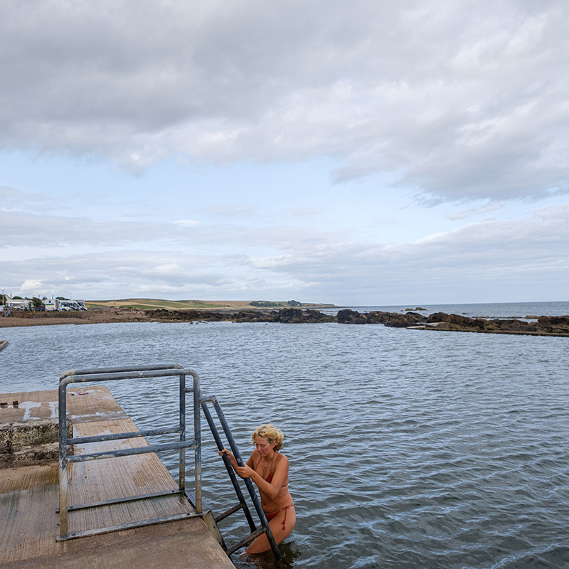 Scottish-seaside-saunas-Cellardyke-03 A person in a swimming costume climbs a metal ladder out of a natural swimming pool by the sea, with cloudy sky and rocky shore in the background.