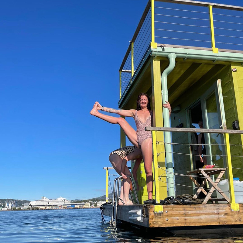 Scottish_Seaside-Saunas-Retreats-02 Two women in swimming costumes stand on a jetty by a yellow houseboat; one performs a standing split, the other stands behind, partially obscured. Water and distant cityscape are visible.