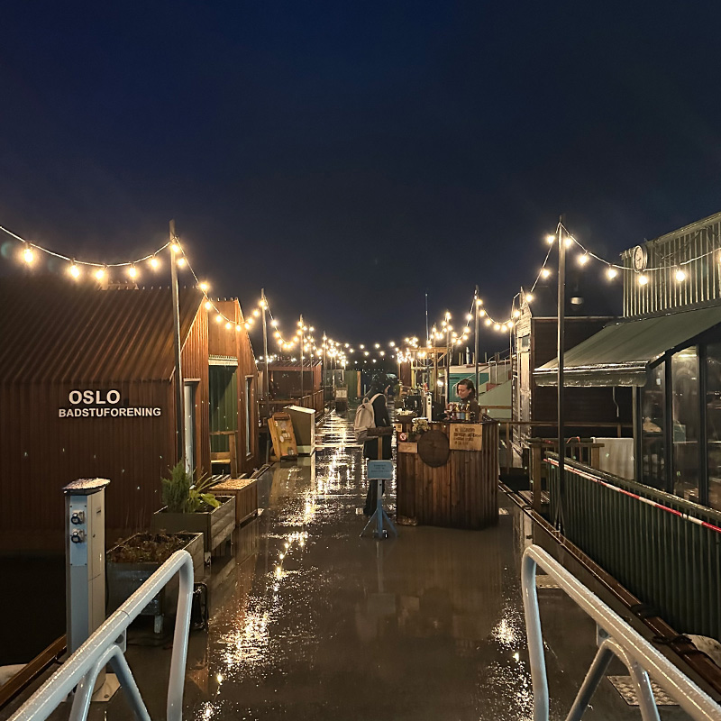 Scottish_Seaside-Saunas-Retreats-03 A wet boardwalk at night with fairy lights overhead, reflecting on the ground. Buildings labelled "Oslo Badstuforening" line the path; one person stands by a wooden stall.