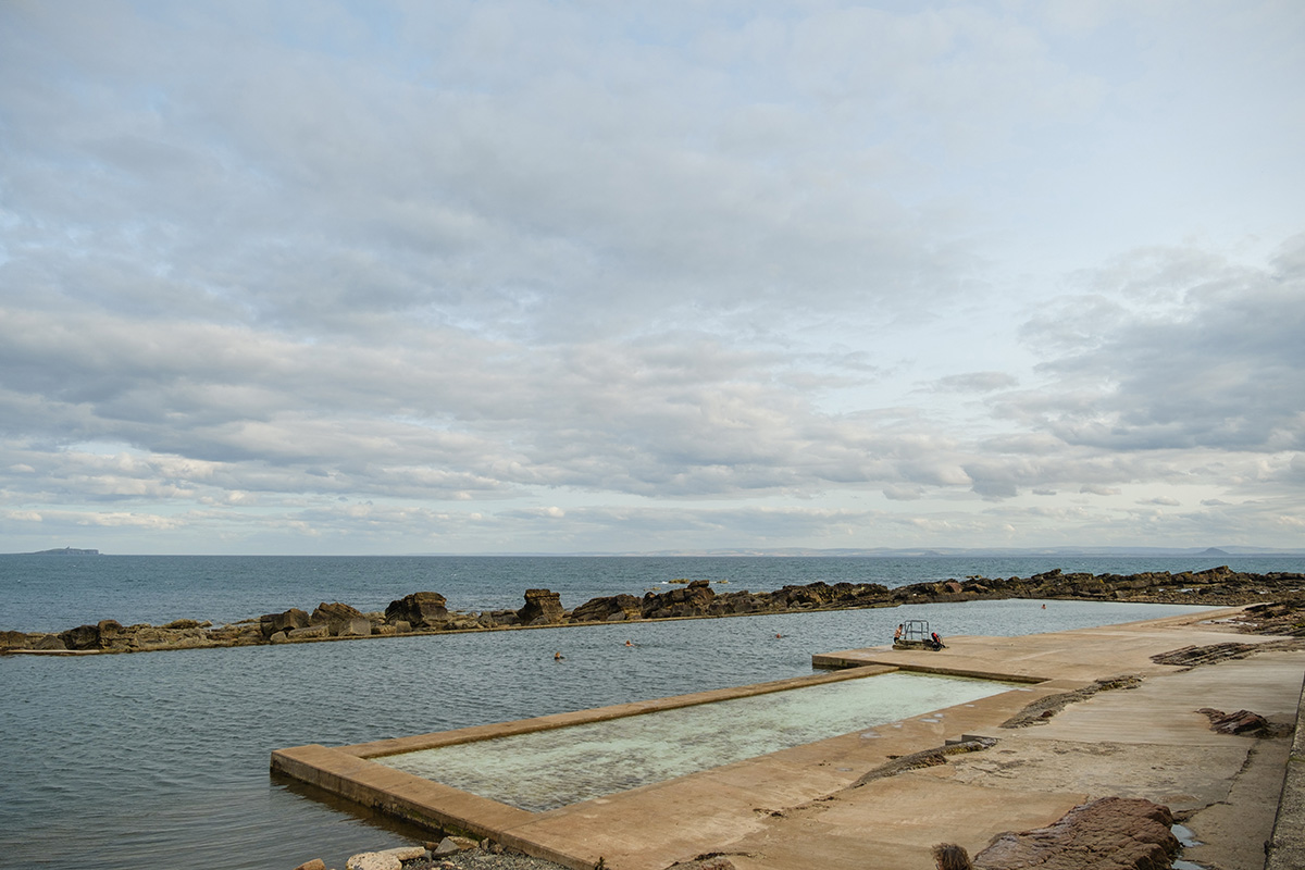 A rectangular ocean pool is built beside a rocky shoreline with calm seawater, under a mostly cloudy sky. A few people are swimming and a lifeguard chair is visible.