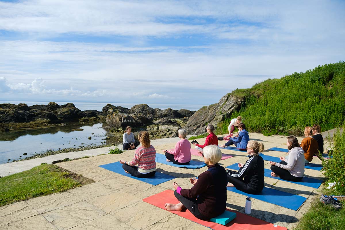 A group of people practise yoga and meditation on mats outdoors near a rocky shoreline and water under a partly cloudy sky.