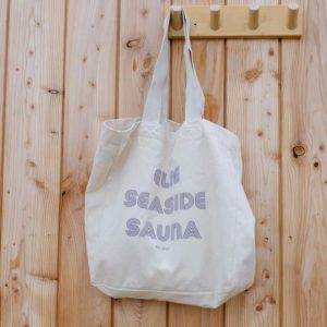A Large Cotton Bag with “ELIE SEASIDE SAUNA” printed on it hangs on a wooden wall hook attached to a light wood panelled wall.