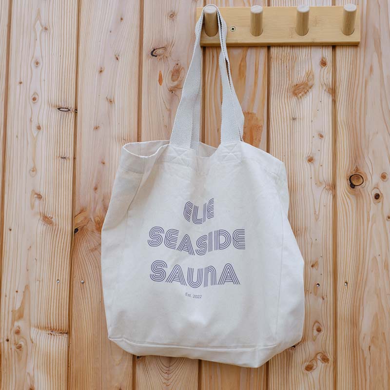 A Large Cotton Bag with “ELIE SEASIDE SAUNA” printed on it hangs on a wooden wall hook attached to a light wood panelled wall.