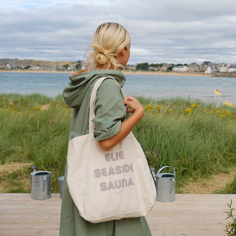 A woman with blonde hair in a green coat stands on a wooden decking by the sea, holding the Large Cotton Bag. Grassy dunes, metal buckets, and water are visible.