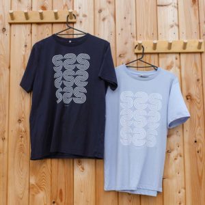 Two men’s basic T-shirts, one navy and one light blue, hang on wooden hooks. Both shirts feature a white geometric “S” pattern on the front.