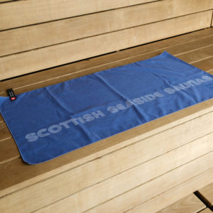 A blue towel with the words “SCOTTISH SEASIDE SAUNAS” printed on it is laid out on a wooden bench.