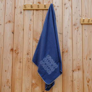 A Quickdry large towel in blue with a white geometric pattern hangs on a wooden peg rail mounted to a light-coloured wooden wall.