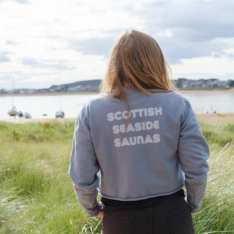 A person stands outdoors facing a body of water, wearing the WOMEN'S BOXY CROPPED SWEATSHIRT with “Scottish Seaside Saunas” printed on the back. Grass and boats appear in the background.
