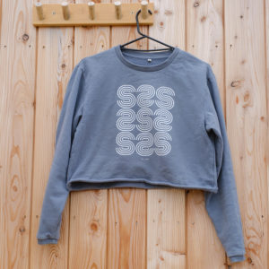 A WOMEN’S BOXY CROPPED SWEATSHIRT in grey, featuring a white geometric “S” pattern, hangs on a black hanger from a wooden hook against a light wood wall.