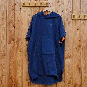 A blue hooded poncho with short sleeves and a large front pocket hangs on a wooden hanger against a wooden wall with coat hooks.