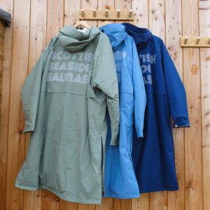 Three EVO waterproof changing robes—in olive green, light blue, and navy—are hanging on hooks attached to a wooden wall. Large printed text is displayed on the back of each robe.