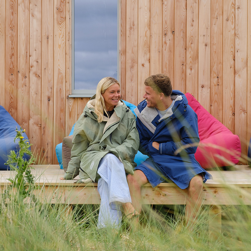 Two people wearing EVO waterproof changing robes sit on a wooden decking in front of a wood-panelled building, smiling at each other, with beanbags and tall grass visible in the scene.
