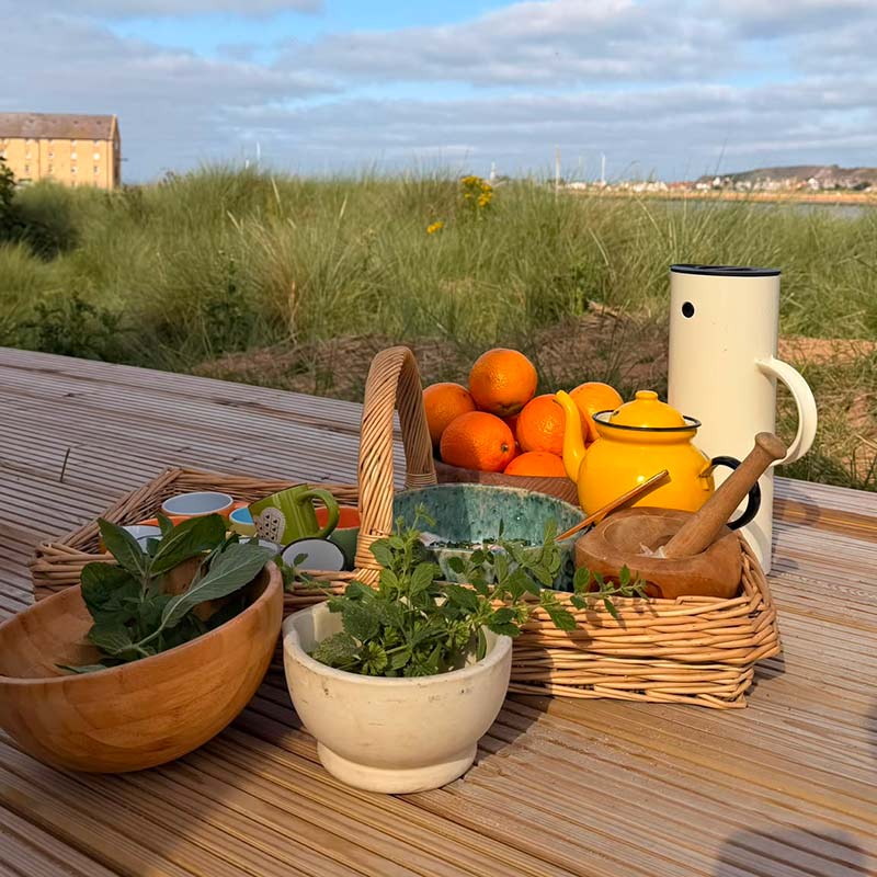 Scottish-Seaside-saunas-events-3 A picnic set-up on a wooden surface with herbs, oranges, a yellow teapot, a white jug, bowls, and a wicker basket, set against grassy dunes and a partly cloudy sky.