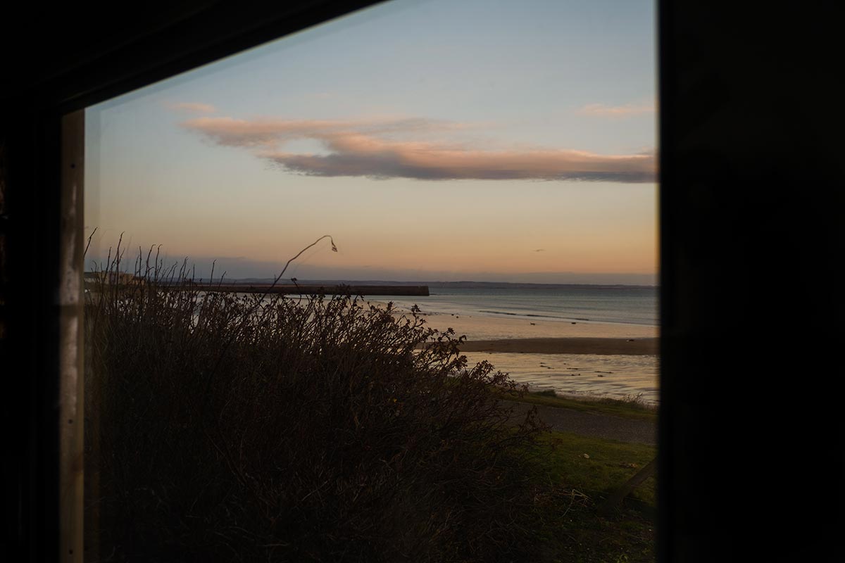 View of a calm beach with low tide and sparse bushes, seen through a window at sunset under a partly cloudy sky.