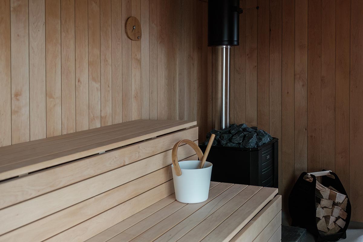 A wooden sauna interior with bench seating, a white bucket and ladle, a heater with stones, and a stack of firewood in the corner.