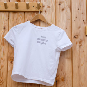 A white T-shirt with “ELIE SEASIDE SAUNA” printed on the front hangs on a wooden hanger against a light wooden wall with hooks.