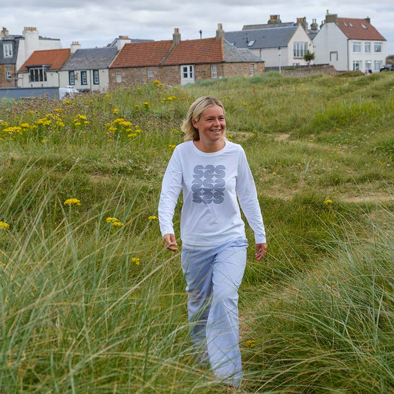 A person wearing the Women’s Long Sleeve T-shirt and light trousers walks through grassy dunes with houses in the background under a cloudy sky.