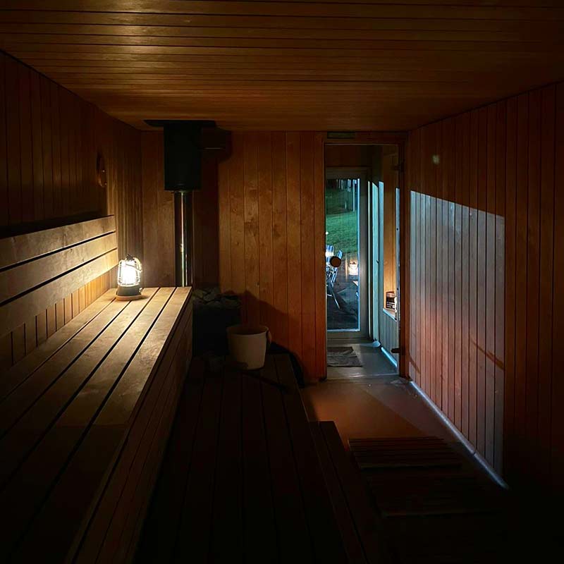 Full-moon-sauna-2 Interior of a wooden sauna with a bench, a lantern, a bucket, and an open door leading outside to a grassy area in low light.