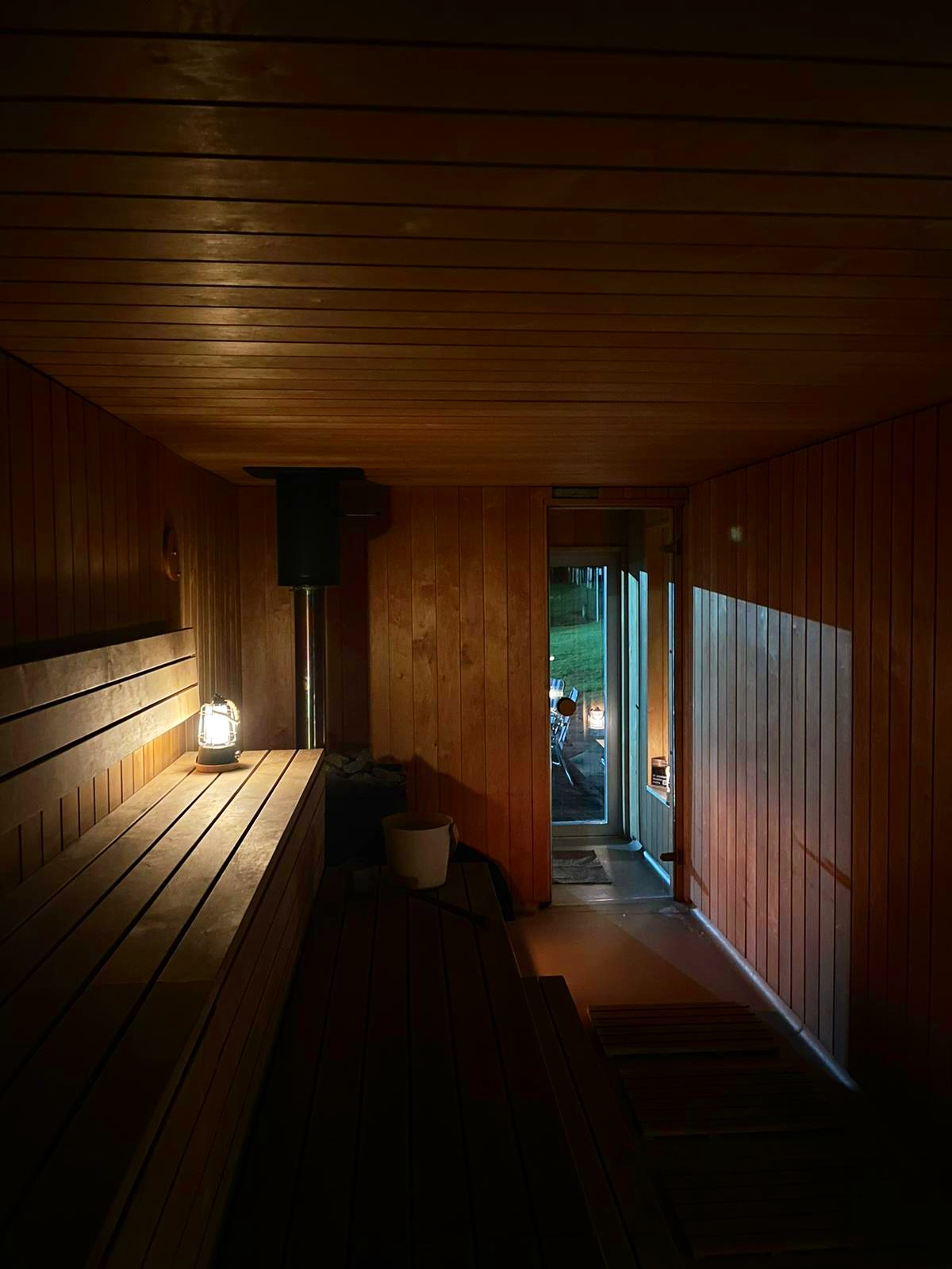 Full-moon-sauna Interior of a wooden sauna with benches and a lantern, door open to the outside at night, light shining in from outside.