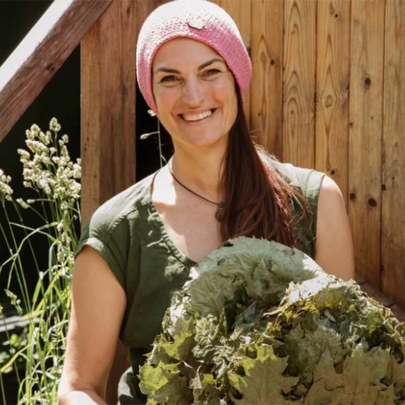 The-Isles-2 A smiling woman wearing a pink knitted beanie holds a large bunch of leafy greens.