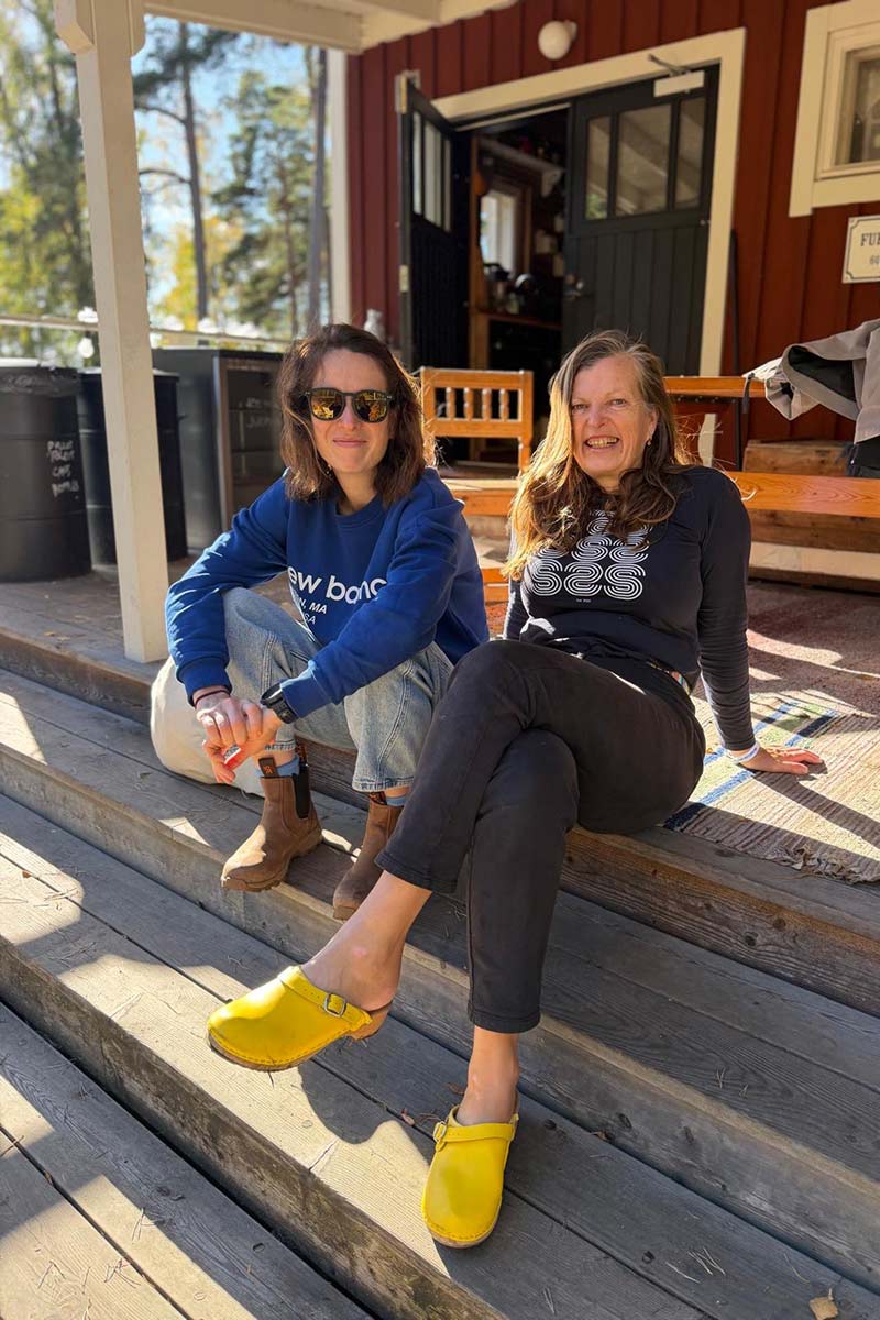 a2551c73-821f-4066-800b-1c250cef8148 Two women sitting on the wooden steps of a building, both wearing sweatshirts and clogs.